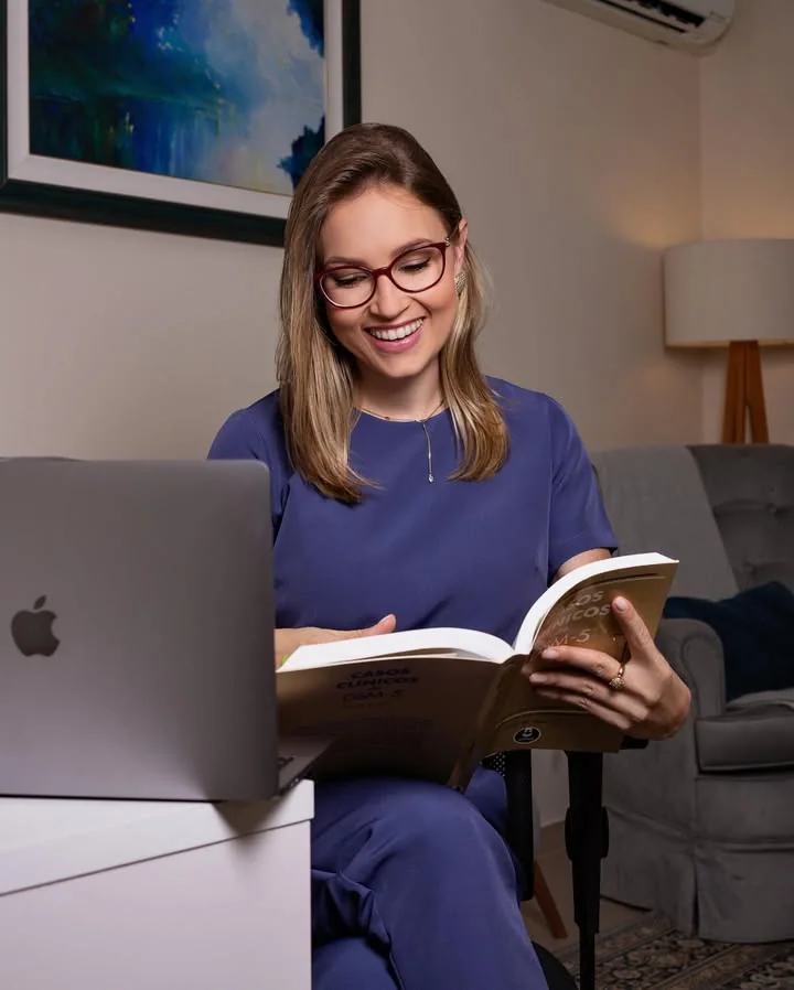 Aline Sá Copetti lendo um livro de casos clínicos no consultório, sorrindo, ao lado de um notebook