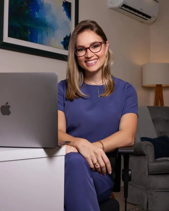 Aline Sá Copetti, psicóloga, sorrindo, sentada no consultório com blusa azul