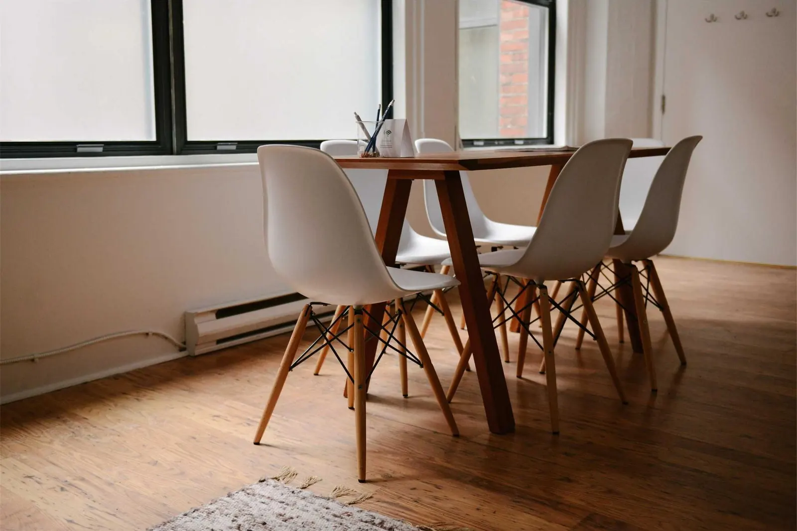 Sala clara com mesa de madeira, cadeiras brancas e janelas amplas